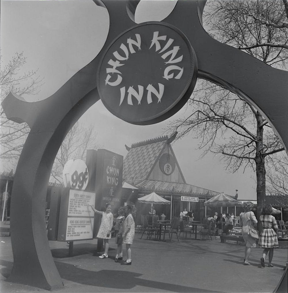 At the New York World's Fair, three young girls look at the ninety-nine cent menu of the Chun King Inn. Customers eat at tables beneath umbrellas.
