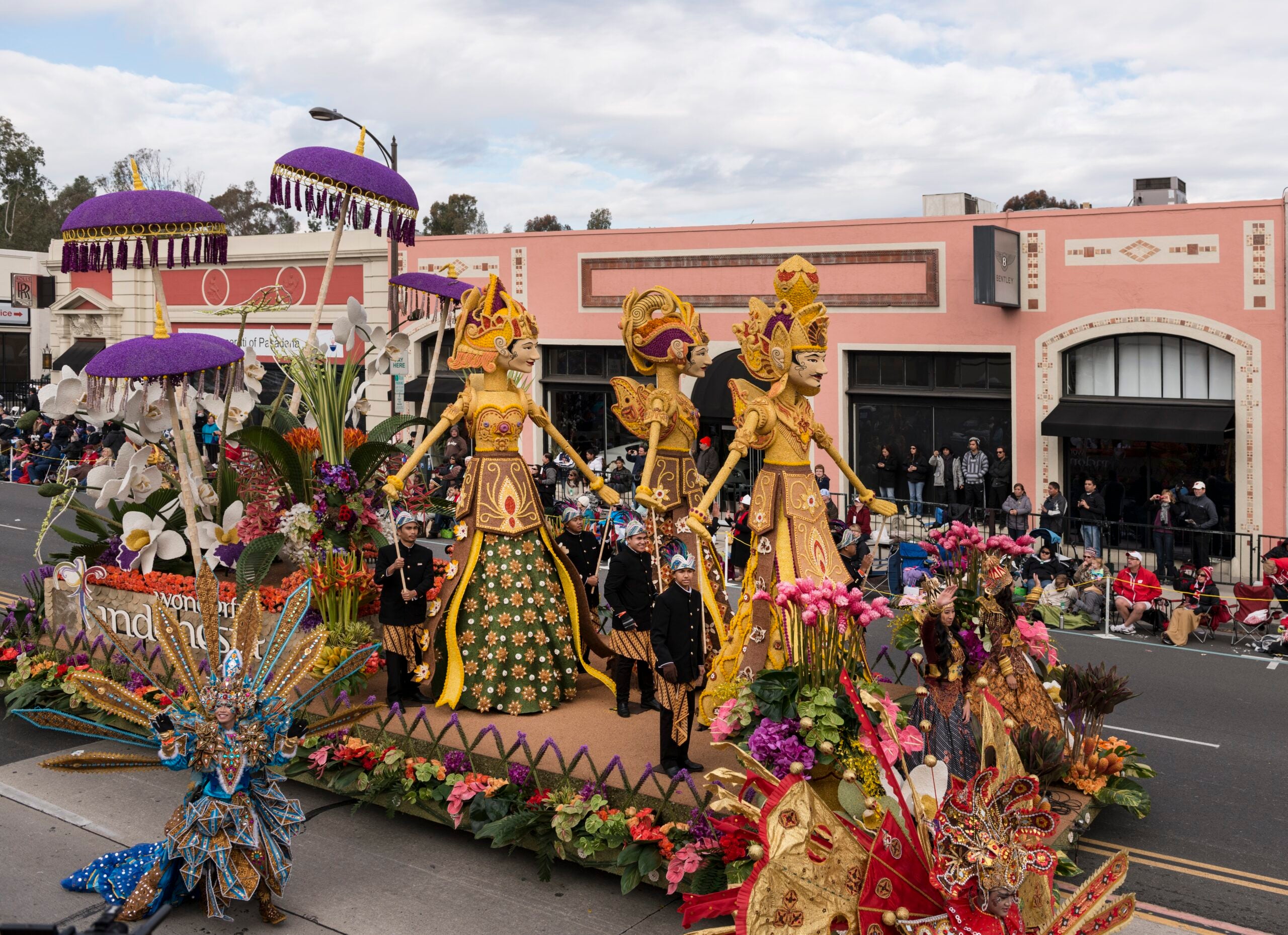 A float with 3 statues in gold costumes, large, purple umbrellas, all surrounded by flowers. Dancers in ornate flower costumes walk next to the float.