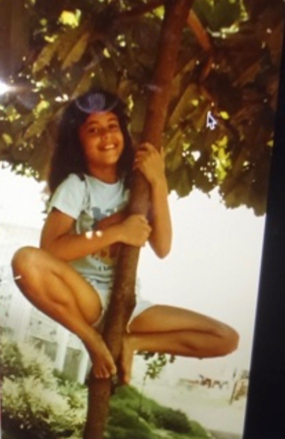 Maya as a young girl wearing shorts and a t-shirt, climbing a tree and smiling.