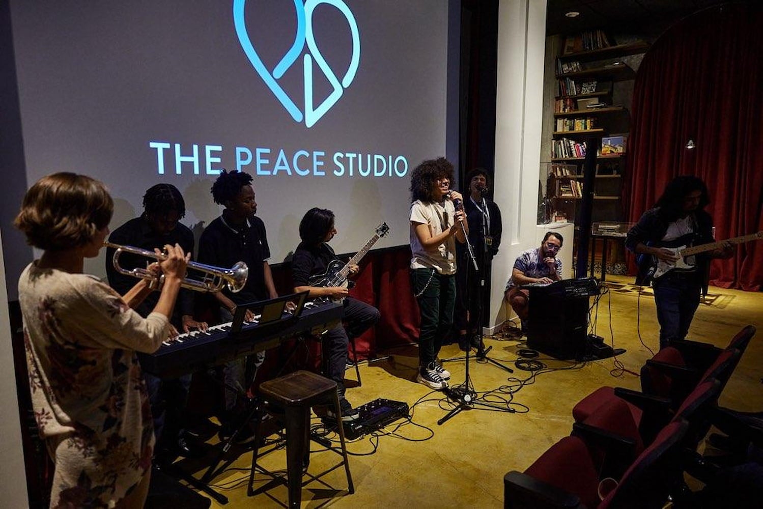 A band plays in front of a screen displaying The Peace Studio. The band has a singer, keyboard, trumpet and guitar players. Books line the wall.