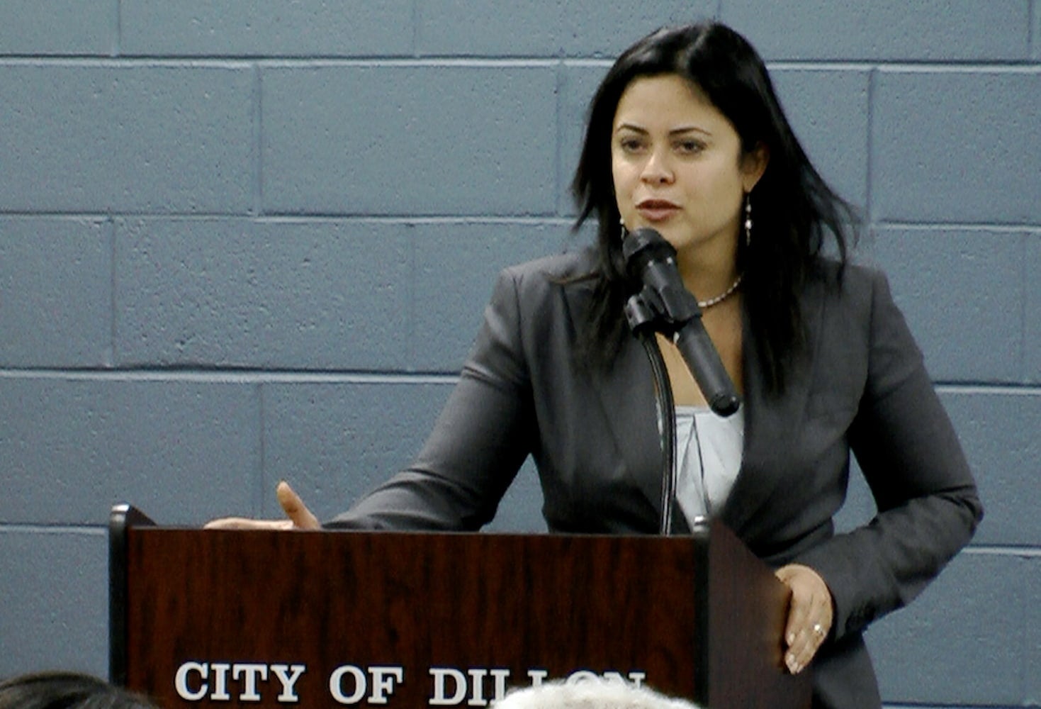 Maya speaks into a microphone at a podium. She is dressed in a business suit.
