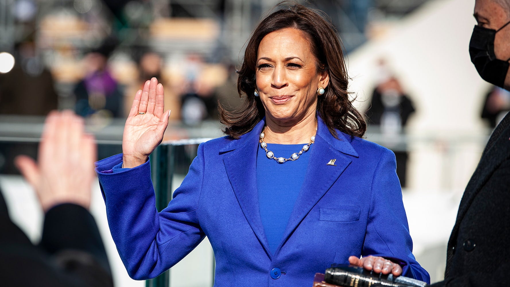 Kamala Harris smiles while being sworn into office. She holds up her right hand, and her left is on a stack of Bibles. She wears a bright blue suit.