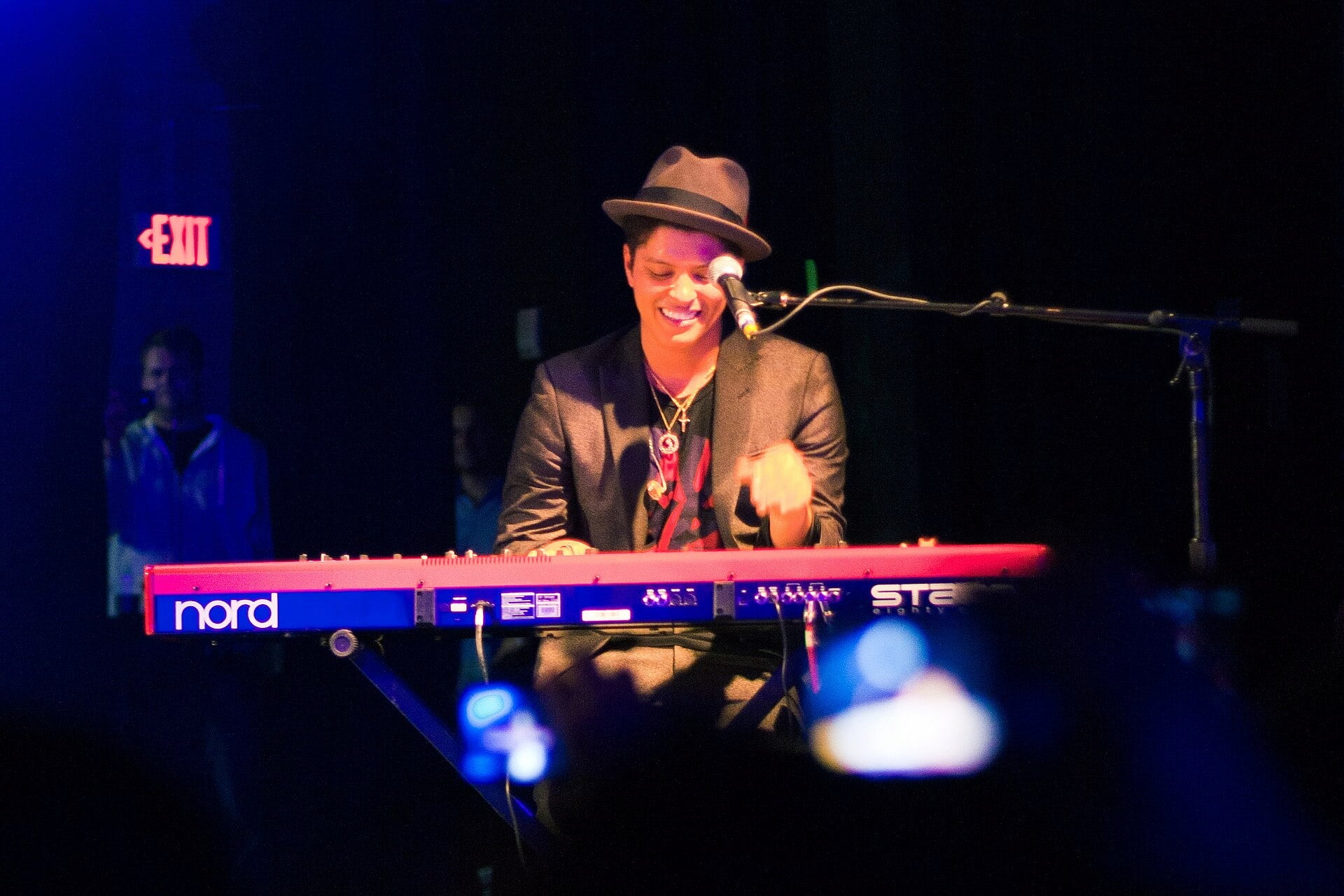 Bruno Mars smiles while playing a keyboard in a darkened room. A microphone is placed in front of his face.