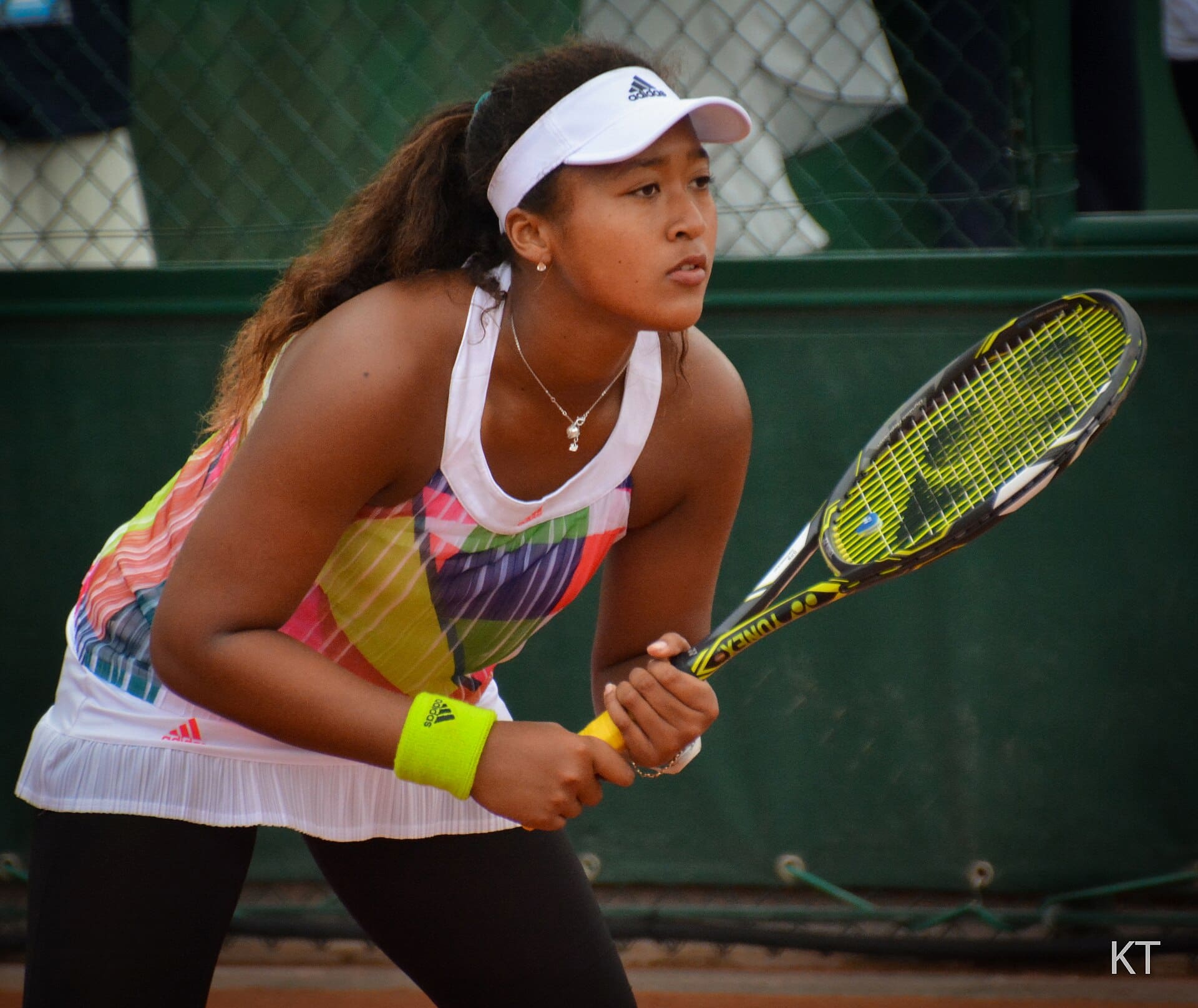 Naomi Osaka crouches and grips her racket with 2 hands, fully focused while waiting for the serve.