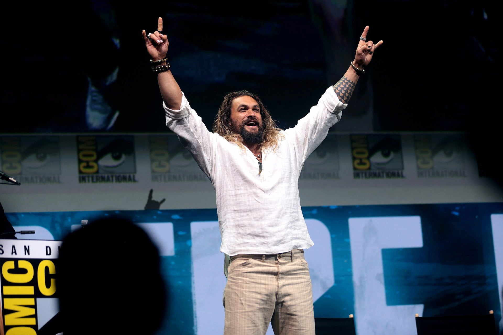Jason Momoa has both arms in the air enthusiastically posing with the horns gesture. Promotional posters for the event paper the background.