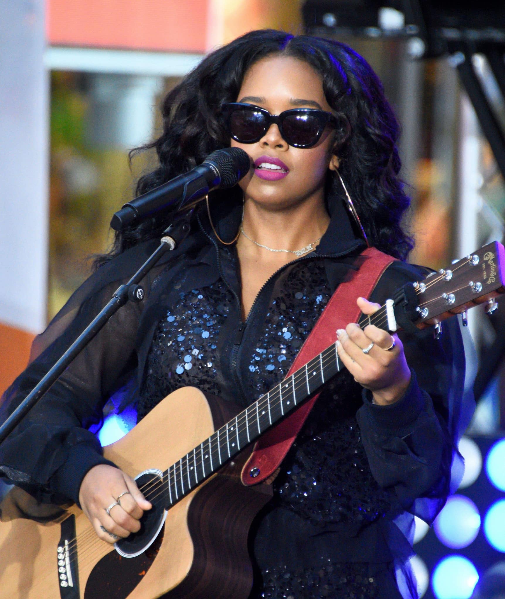 Gabriella Wilson (aka H.E.R.) on stage with a guitar and a microphone. She wears dark sunglasses, large hoop earrings, and a black sequins outfit.