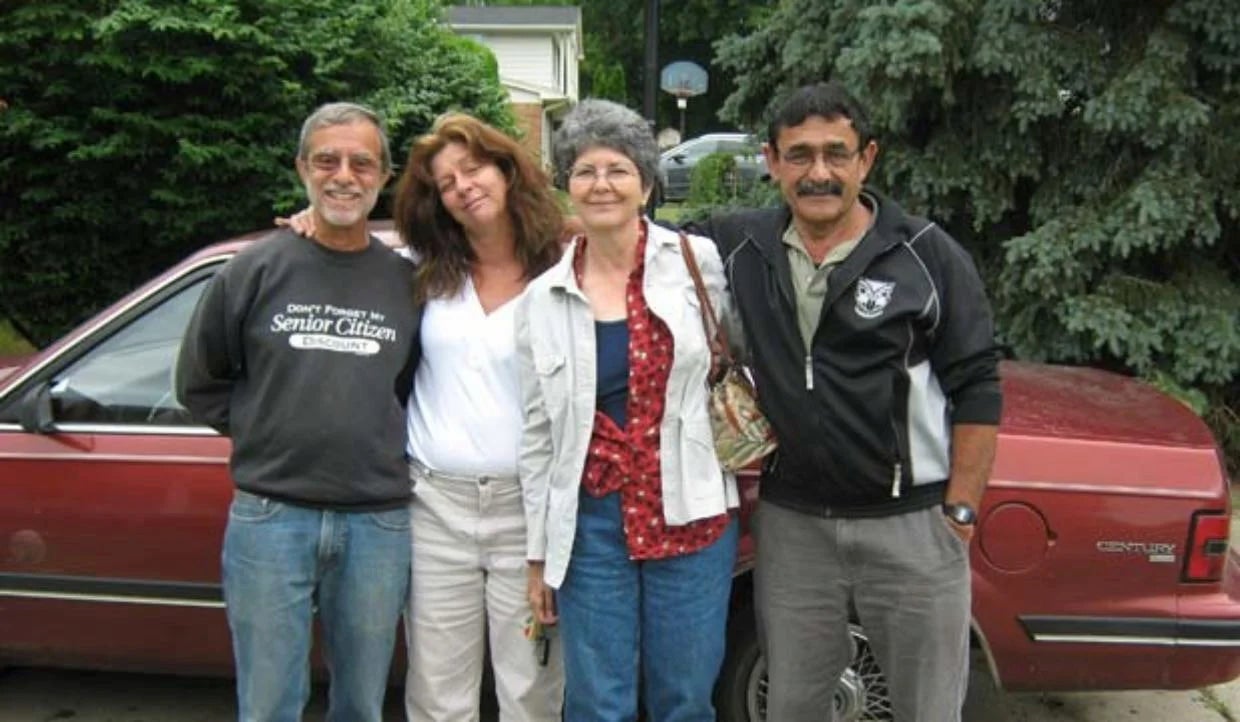 Arthur Beren poses with his family in a driveway. A red sedan, trees and a house show in the background.