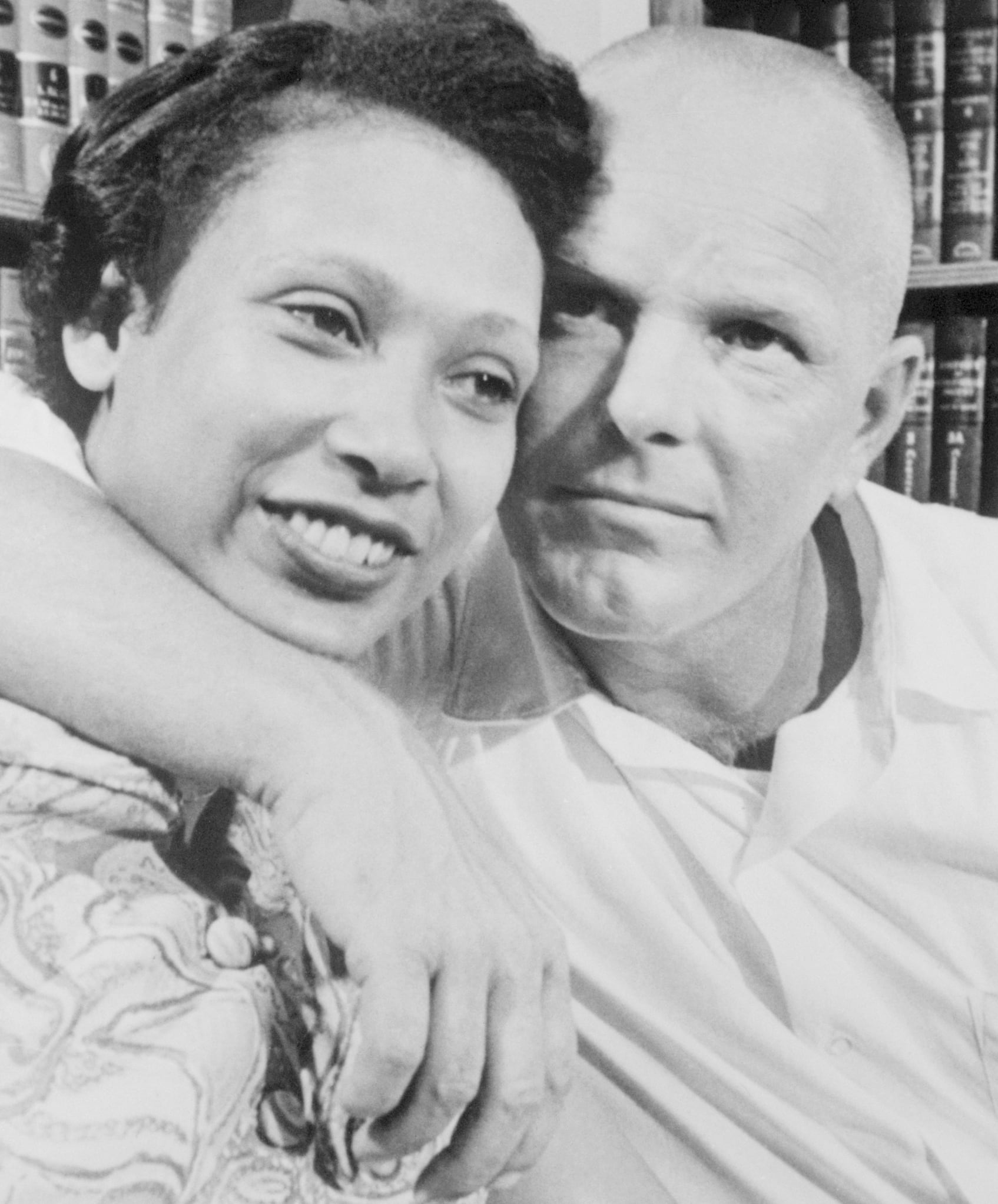 Black and white photo: Mildred smiles as Richard puts his arm around her neck, holding their faces close together. Law books line the shelves behind them.