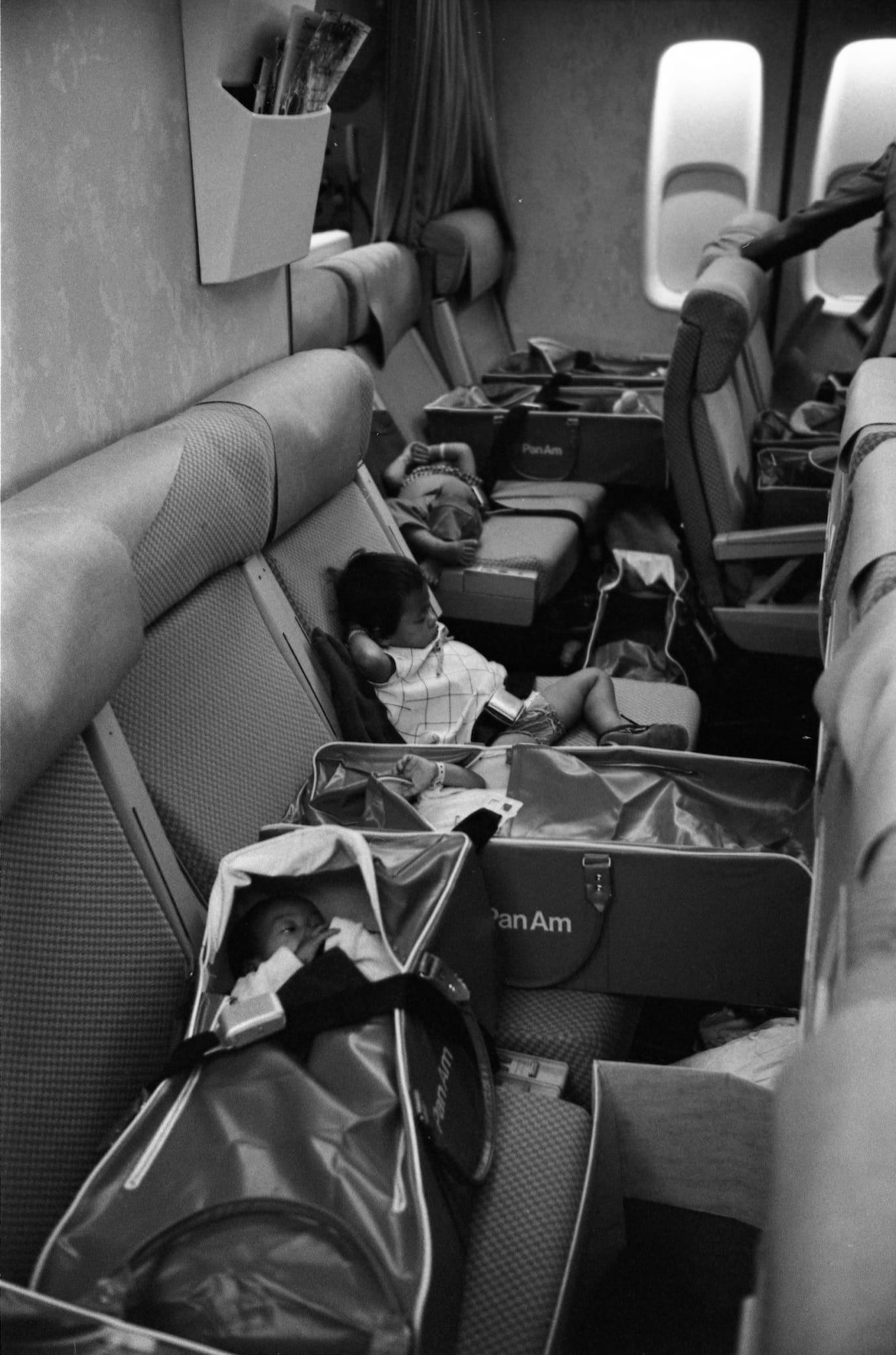 Black and white photo: The back rows of an airplane have a line of babies in travel cribs. One toddler tries to sleep among the infants.