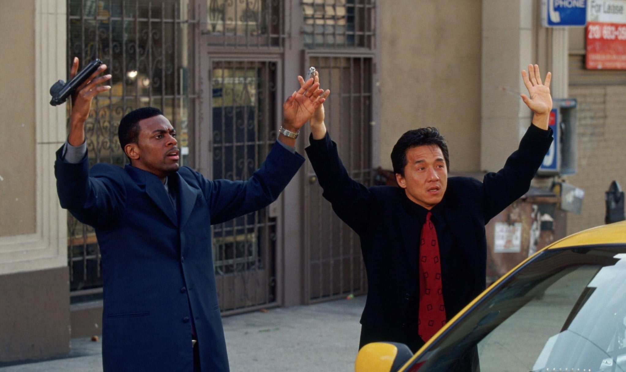 Clip from film Rush Hour: Chris Tucker and Jackie Chan stand on a sidewalk with their hands in the air. Tucker dangles a firearm.