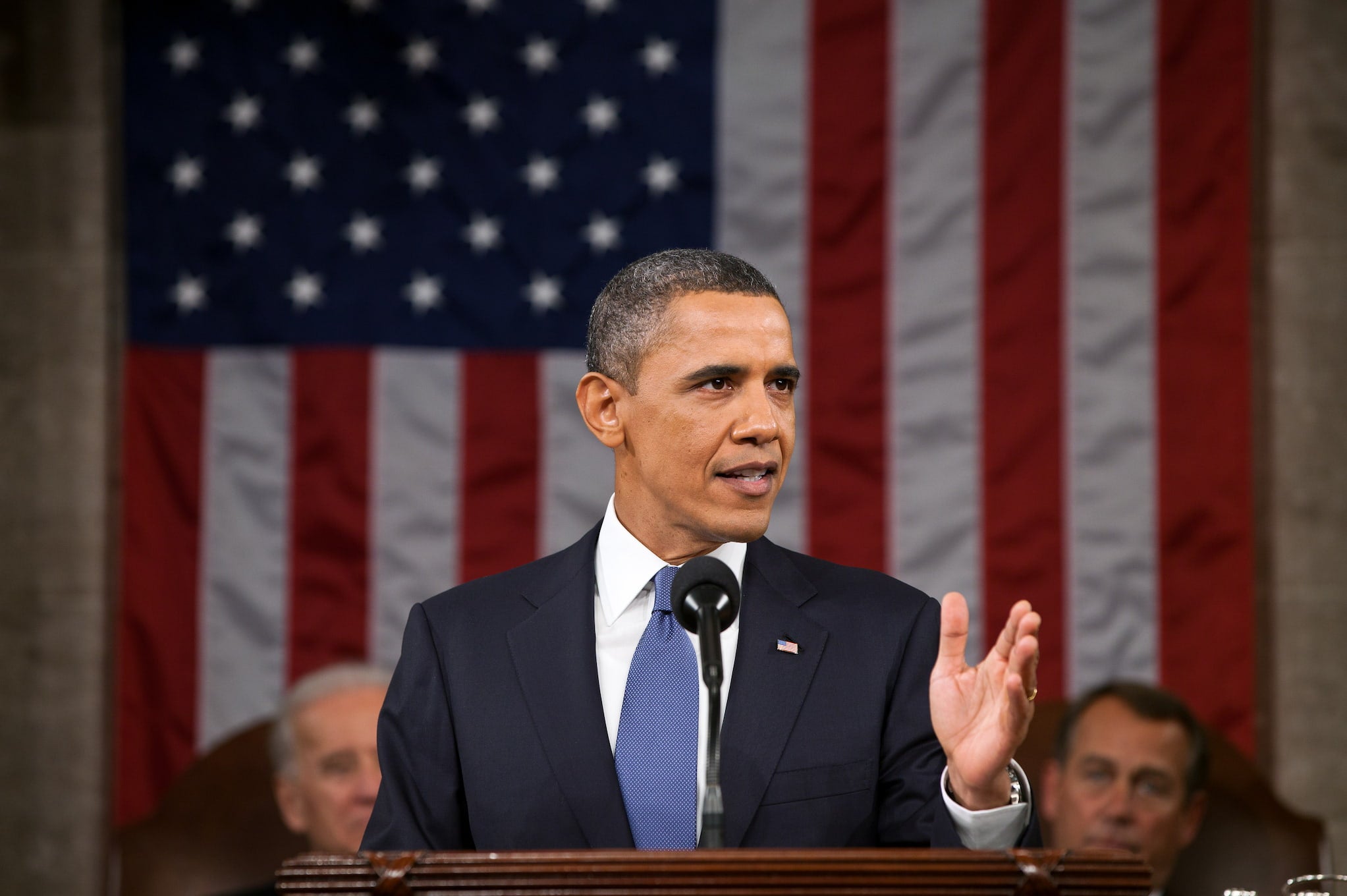 President Obama standing at a podium delivering the State of the Union. The American flag drapes the background.