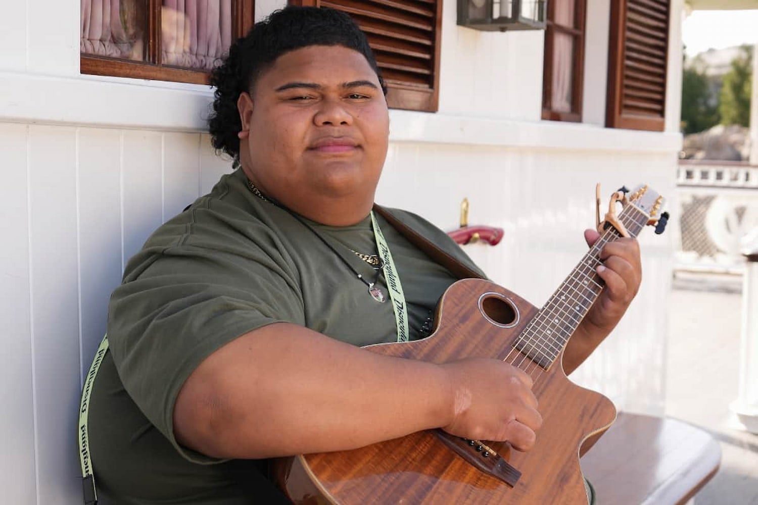Iam Tongi leans against a white house posed to play his guitar. His contemplative expression shows a slight closed-mouth smile.