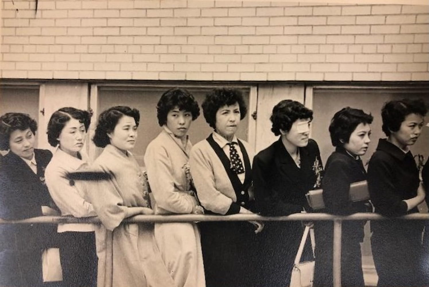Eight Japanese women, nicknamed the Hiroshima Maidens, stand alongside a railing in front of a brick building.