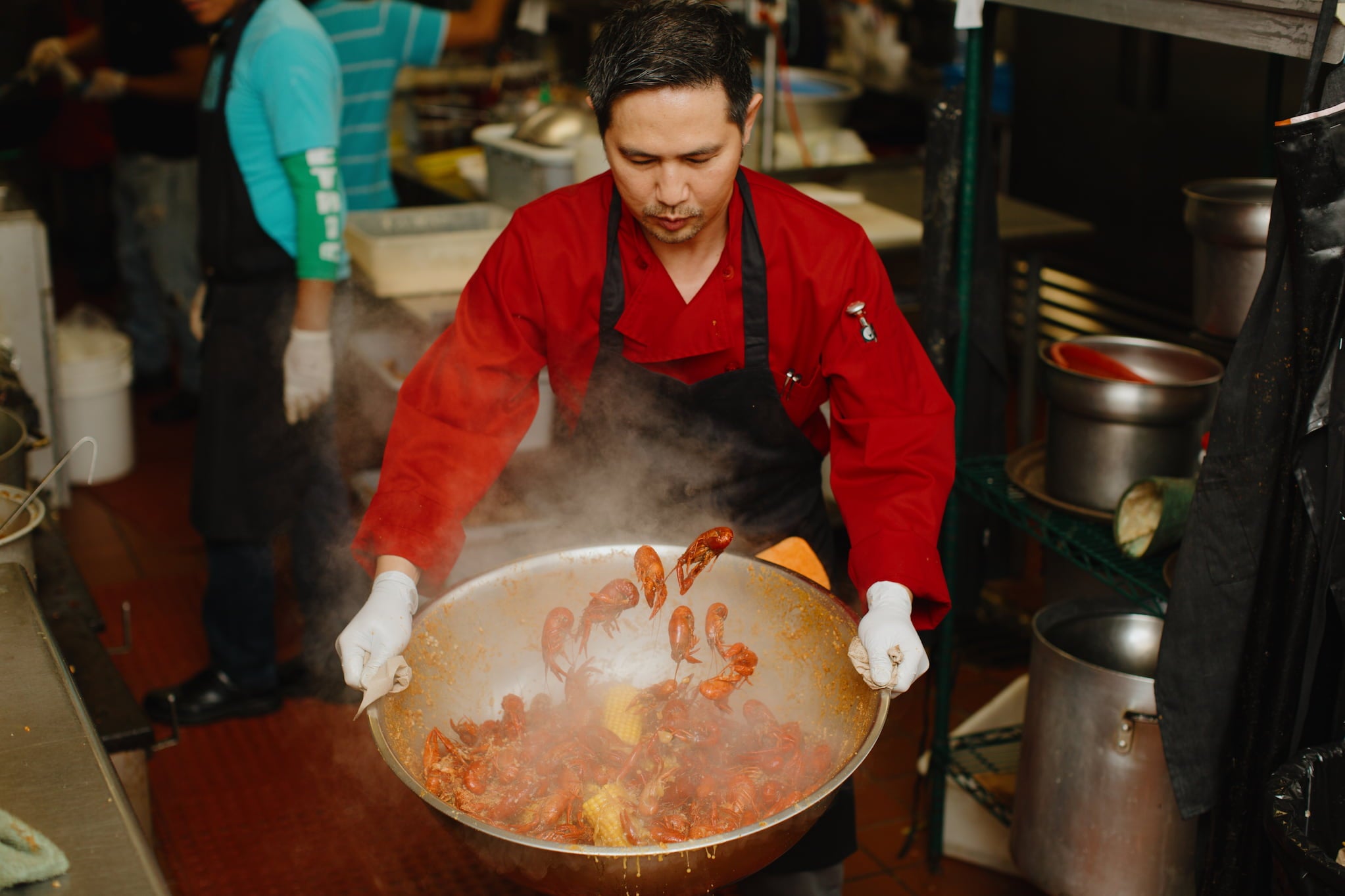 A chef in a red chef coat and black apron holds a giant steaming bowl of crawfish.