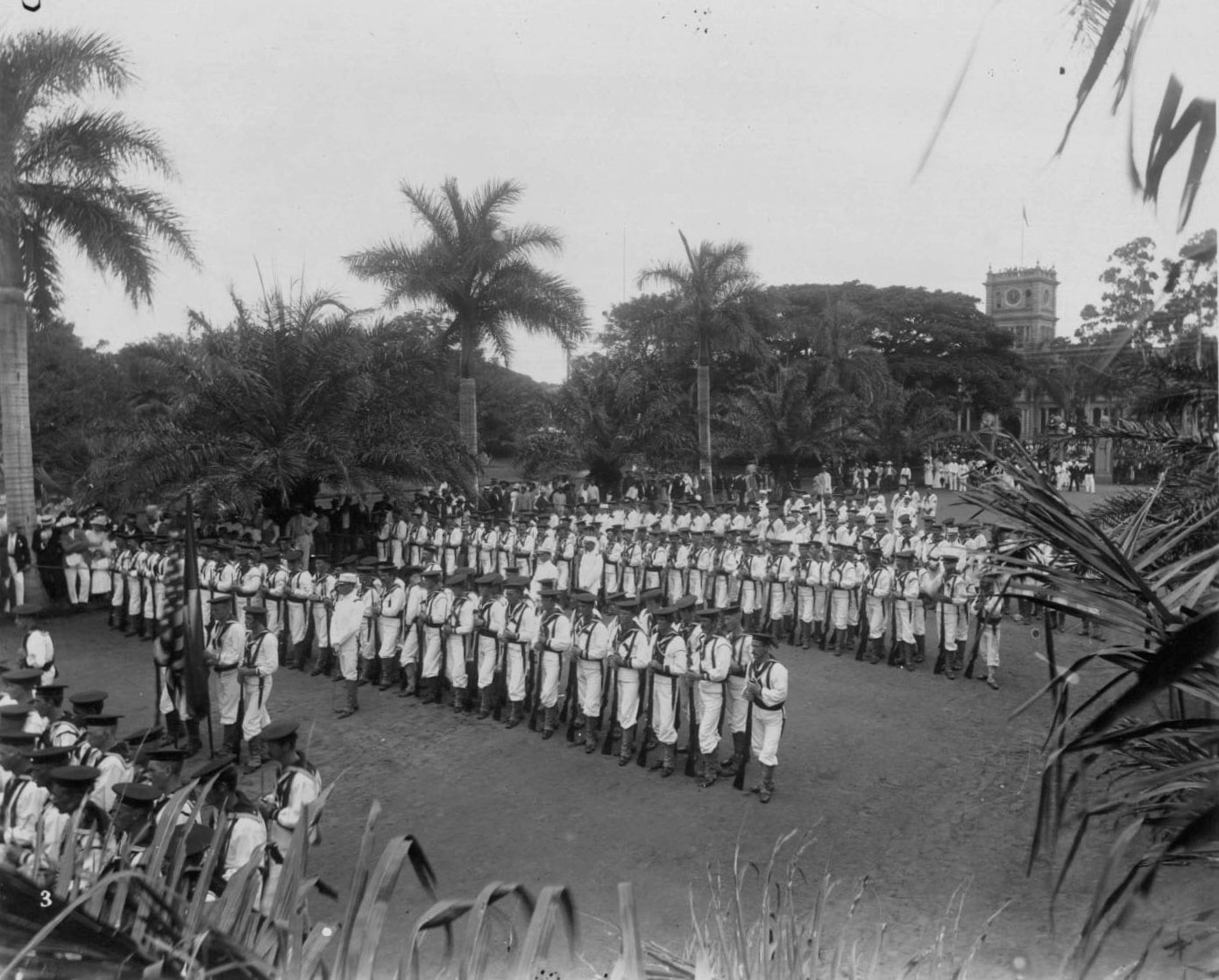 Rows of U.S. Marines, dressed in white uniforms, stand at parade rest with rifles near ‘Iolani Palace. Color guard stand in front.