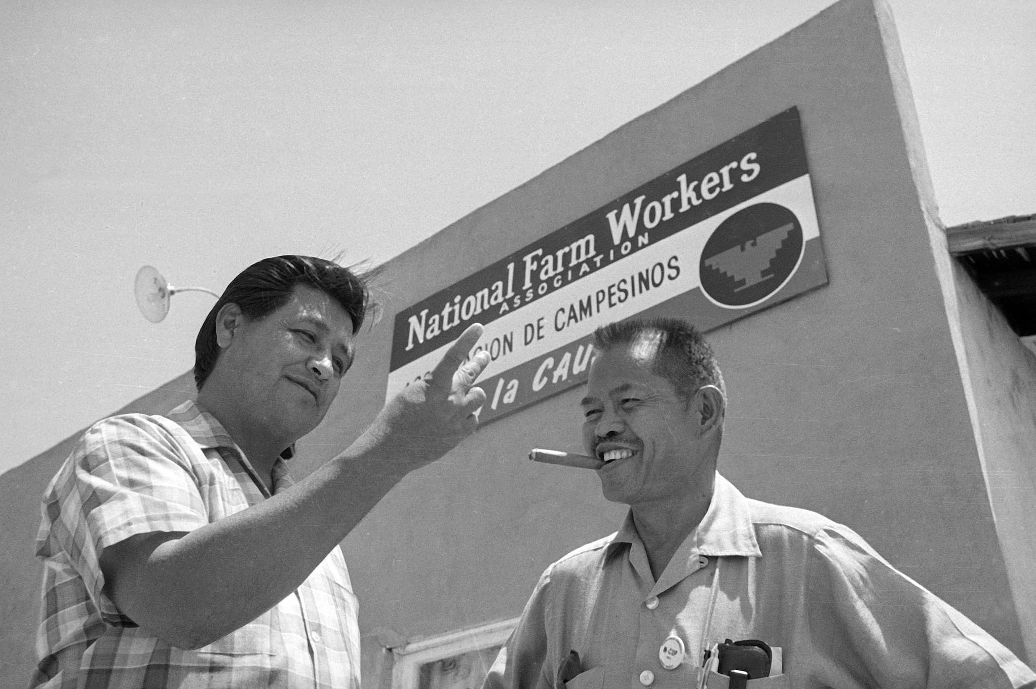 Filipino American Larry Itliong, next to Mexican American Cesar Chavez, stands outside National Farm Workers Association headquarters. Chavez gestures with his hand.