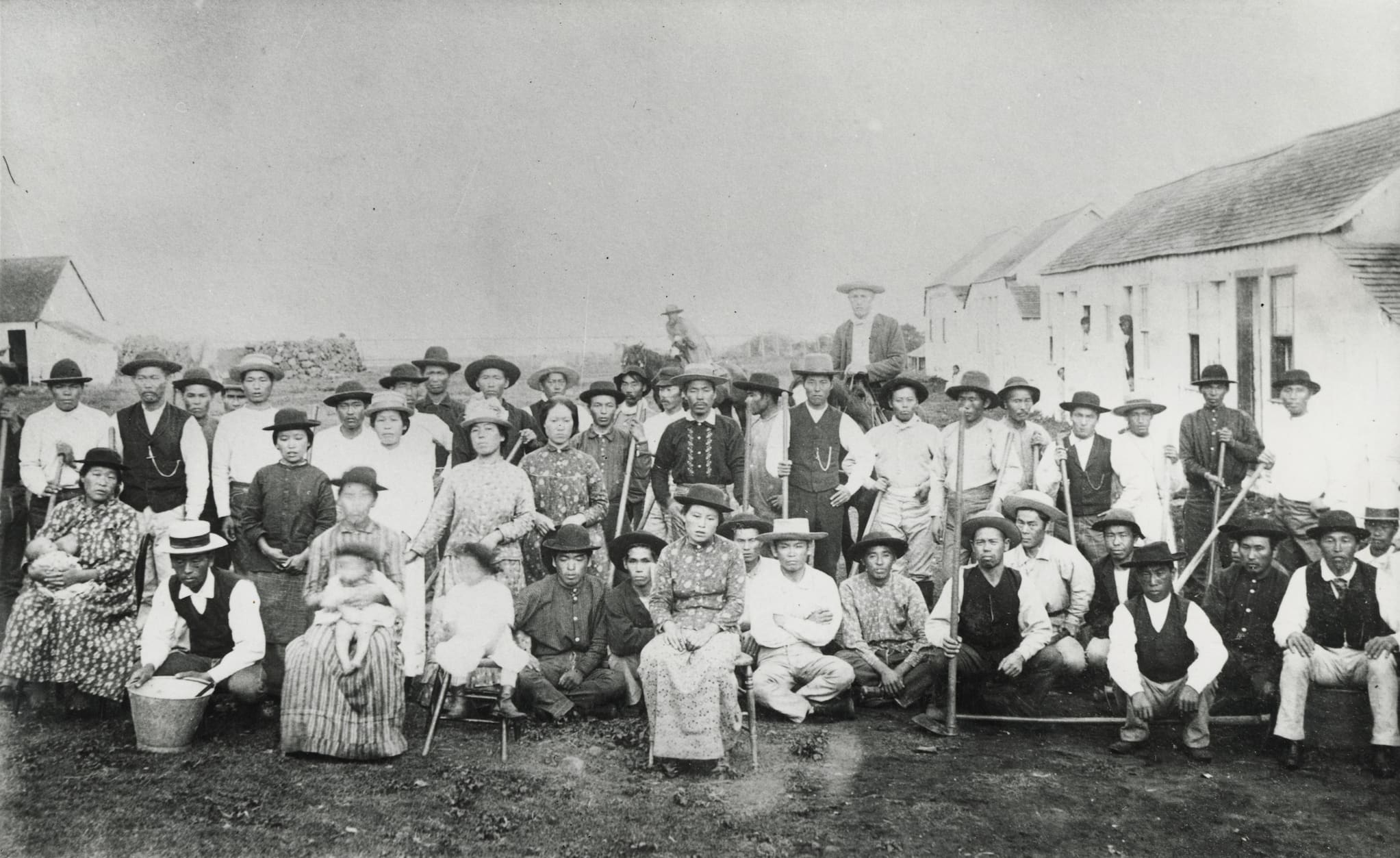 Group of Japanese sugarcane plantation workers and their families, wearing hats and either dresses or pants, vests, long sleeved shirts, pose outside.