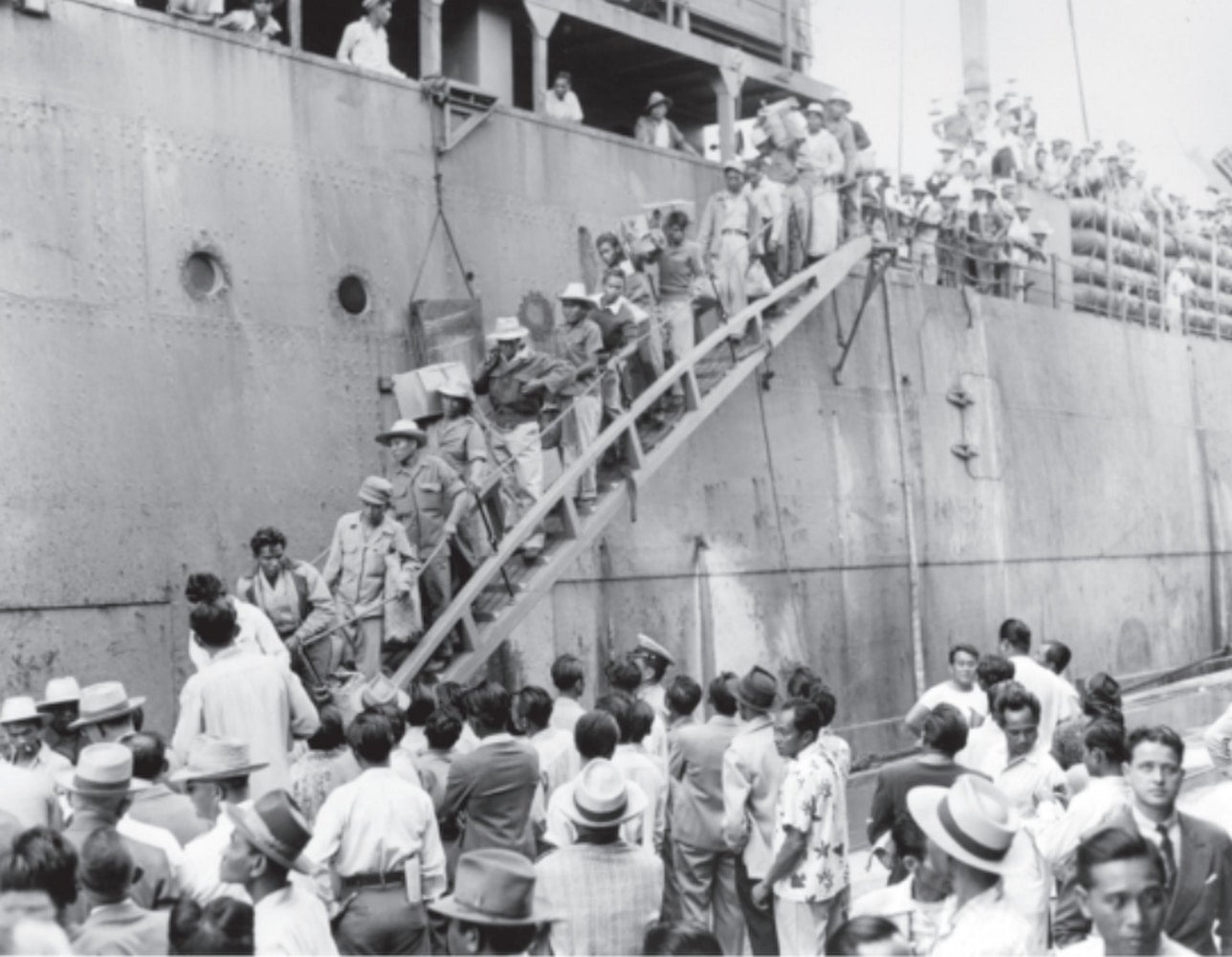 Filipino immigrants line up and walk down the gangway of a ship while holding their belongings. A crowd stands on the dock watching them disembark.