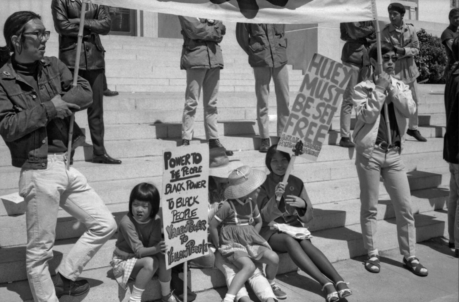 Black and white photo: Vicci Wong and two other girls sit on a curb while holding signs in support of Huey Newton. Other people with signs stand nearby.