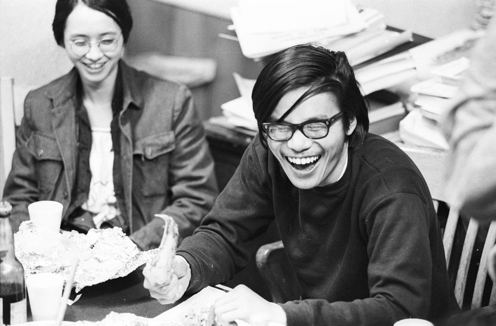 Harvey Dong, a young Chinese American man wearing glasses with thick frames and a dark colored sweater, laughs while seated at table.