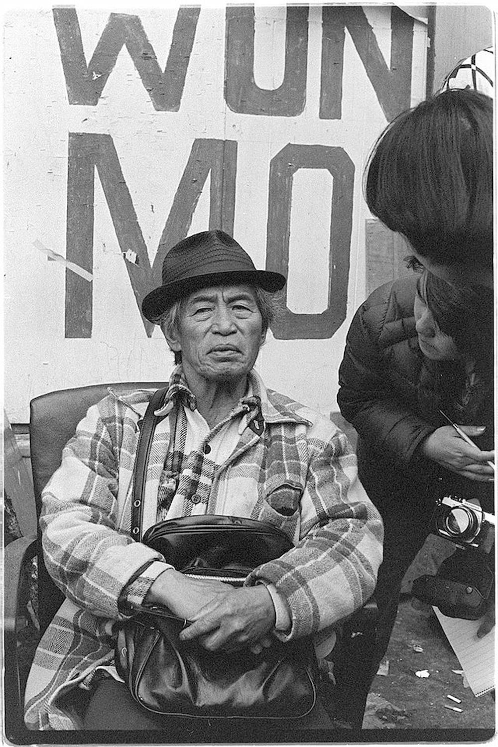 Wahat Tompao, an elderly I-Hotel tenant dressed in a plaid jacket and wearing brimmed hat, sits in a chair outside of the hotel.