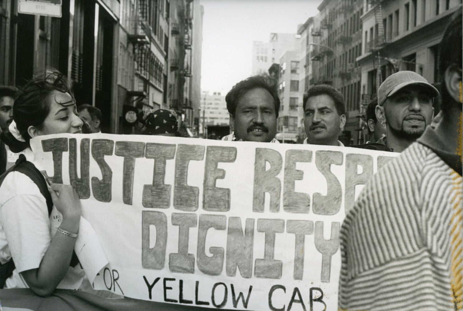 Lease Drivers Coalition members gather in street for protest. Several people stand behind banner reading "Justice. Respect. Dignity."