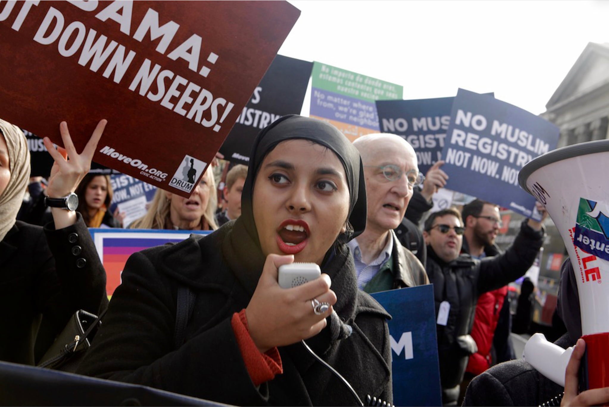 Woman in hijab speaks into a megaphone surrounded by protestors holding a variety of protest signs.