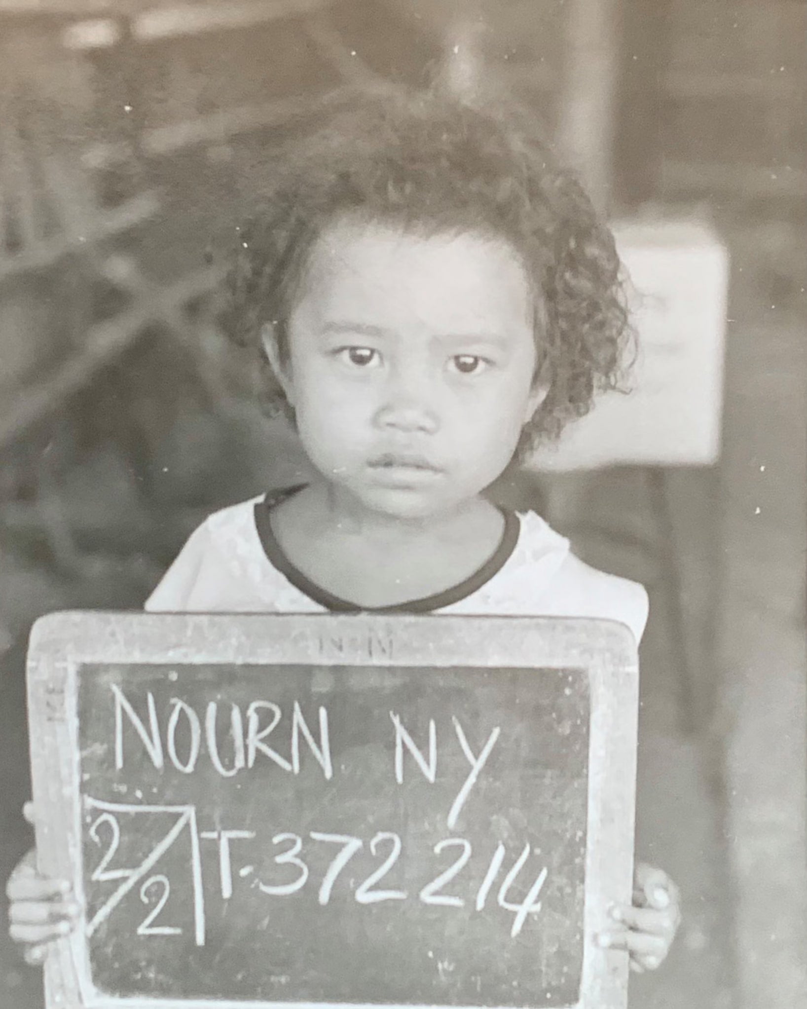 Black and white photo of Ny Nourn as a small child holding up a small chalkboard with her name and a number. Her expression shows concern and fatigue.