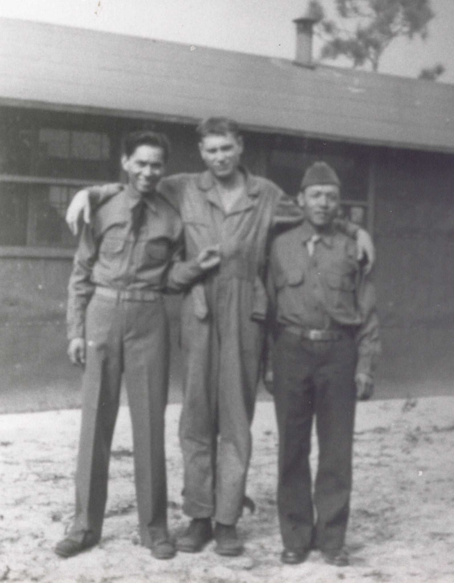 Three men, dressed in U.S. military attire, pose for group photo outside of building. The man in the middle rests his arms over their shoulders.