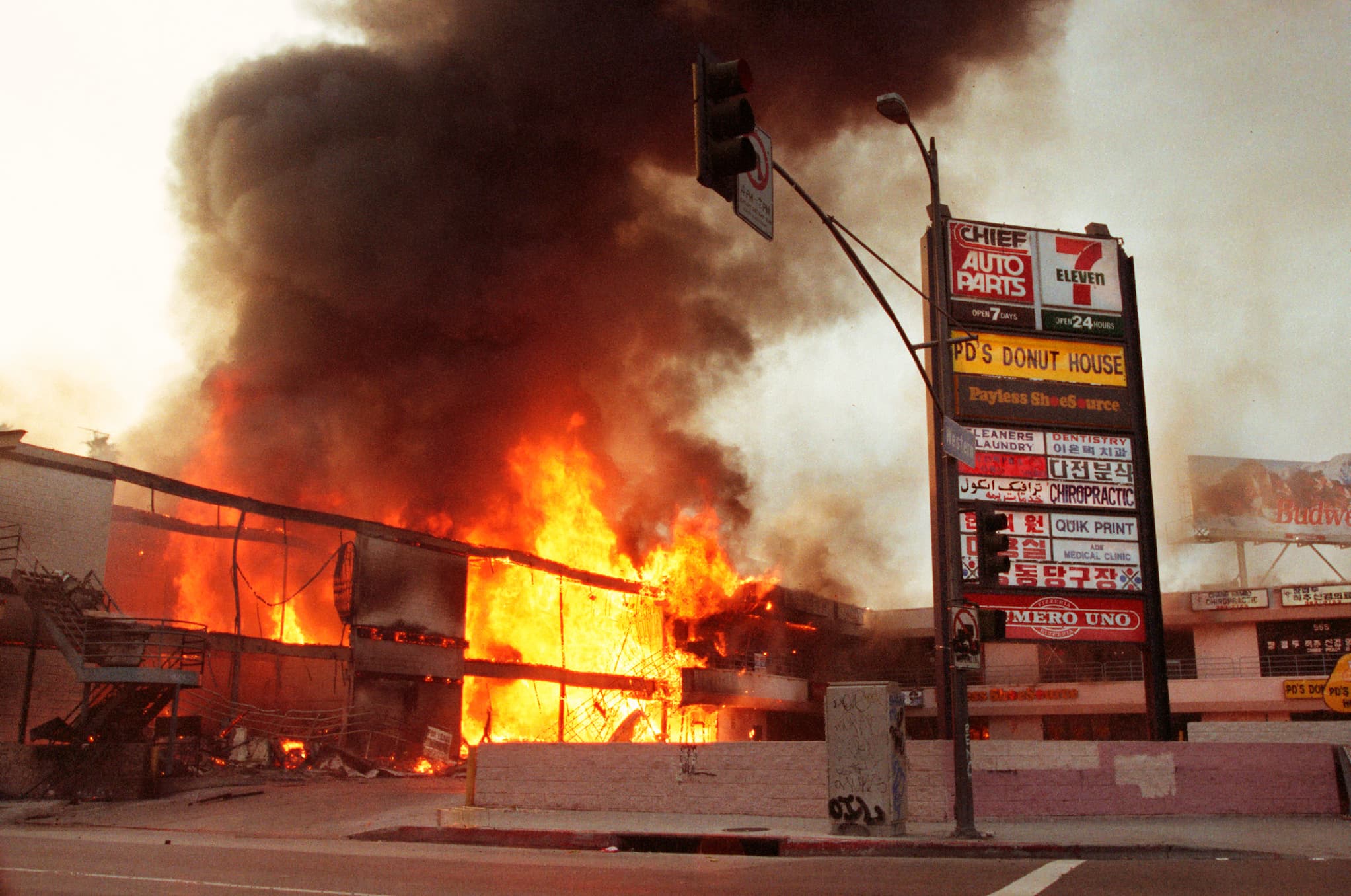 A shopping mall on the left billows out black smoke as it is engulfed in flames. A multi-tenant sign on the right displays business names.