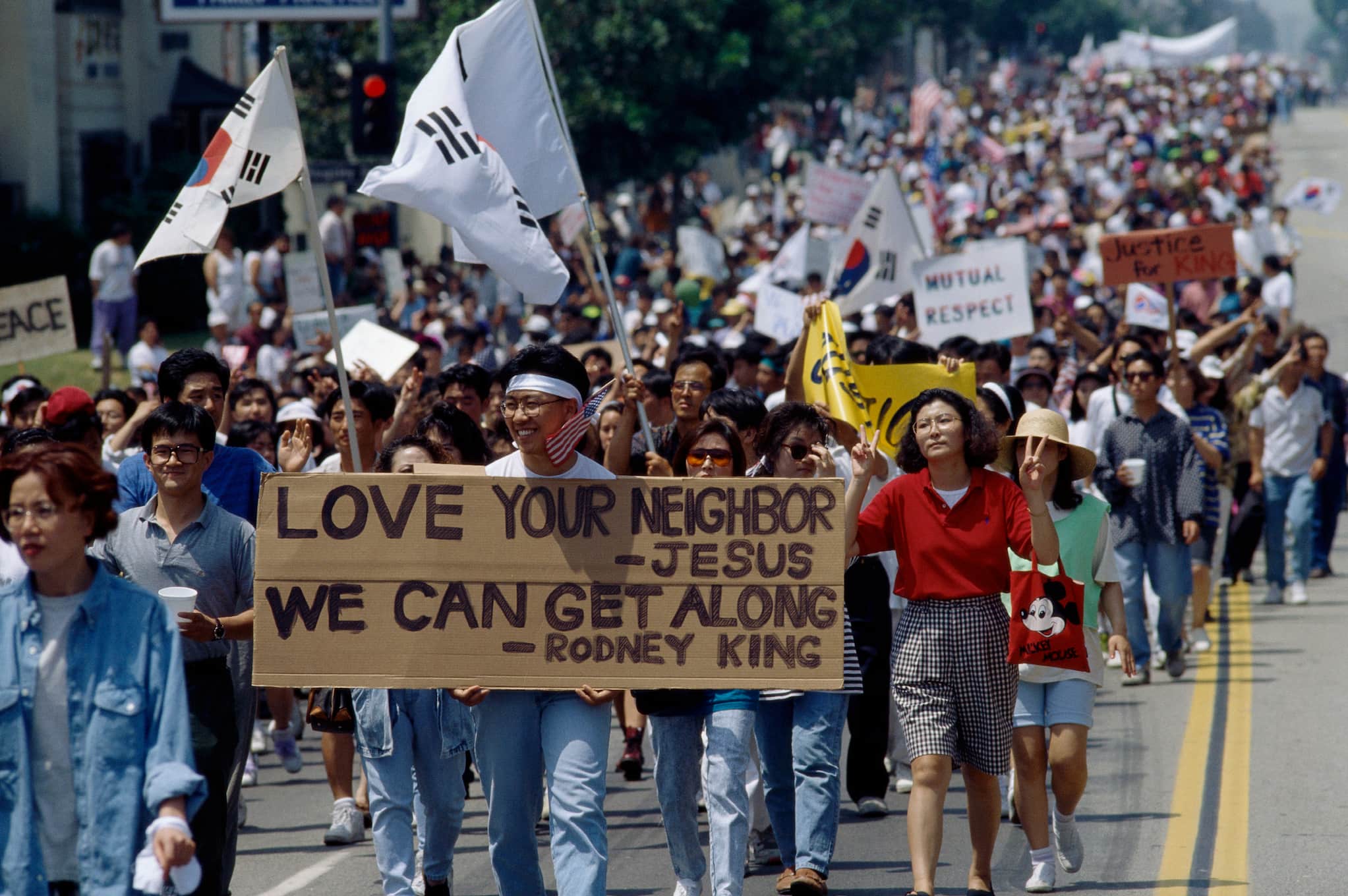 A large group of people walk down the middle of street during peace rally. People hold the flag of South Korea and signs promoting peace.