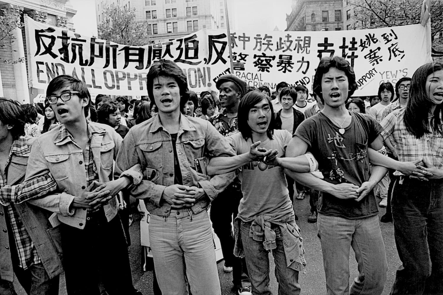 Three Asian American men stand with their arms interlocked. Behind them a crowd of protestors hold up a large banner above them.