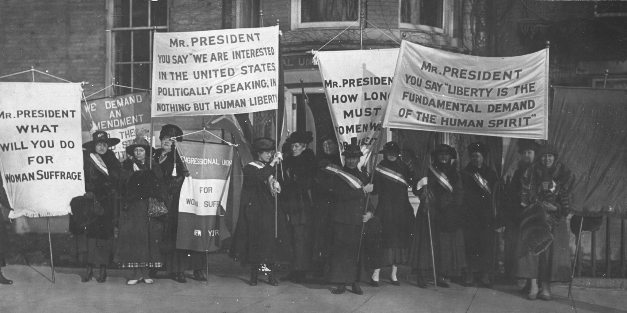 Suffragists, dressed in black dresses, large hats and sashes, hold banners in support of women's suffrage as they stand outside the Cameron House.