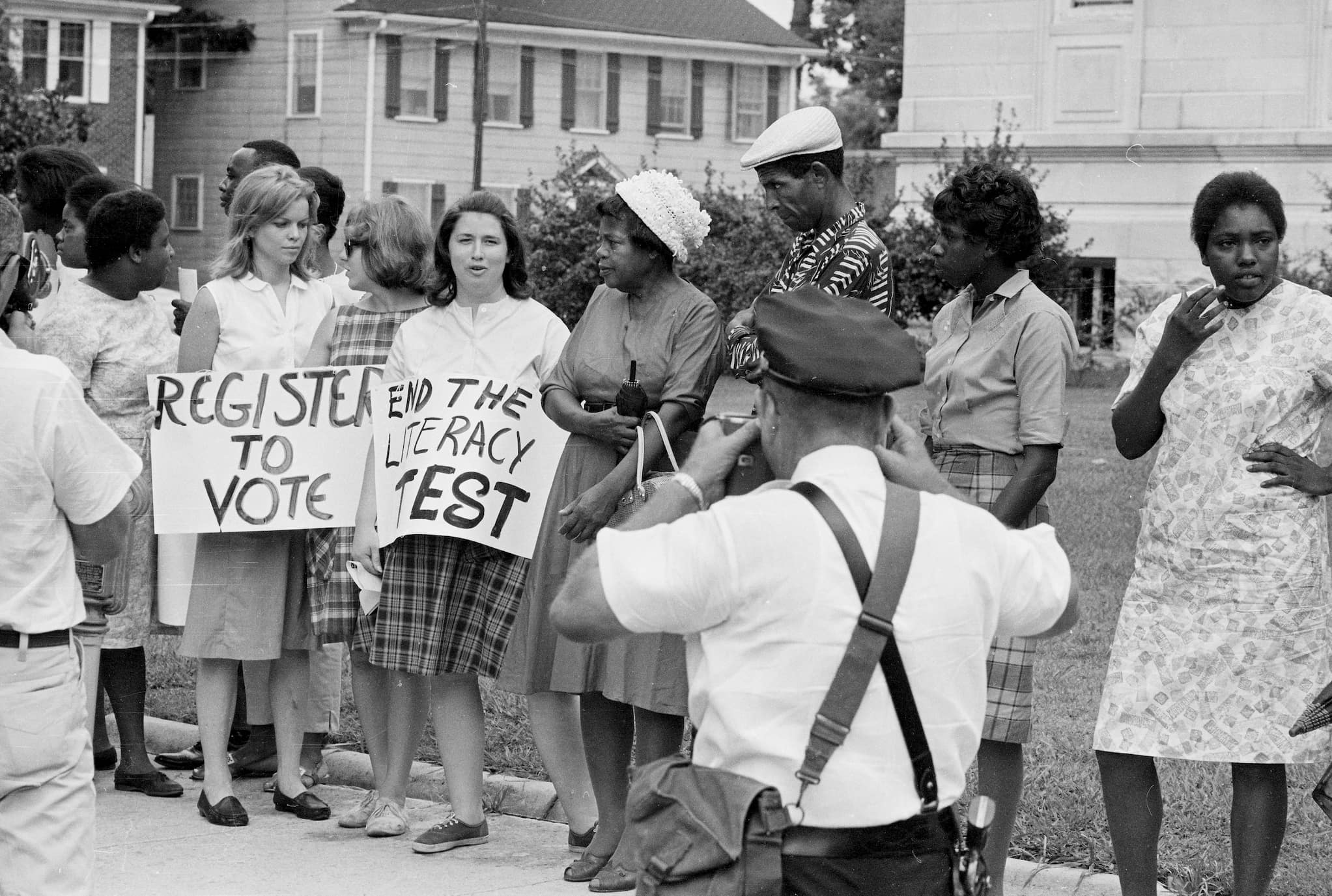 Three white civil rights workers, holding signs related to voter registration, stand with a group of Black citizens gathered on the sidewalk.
