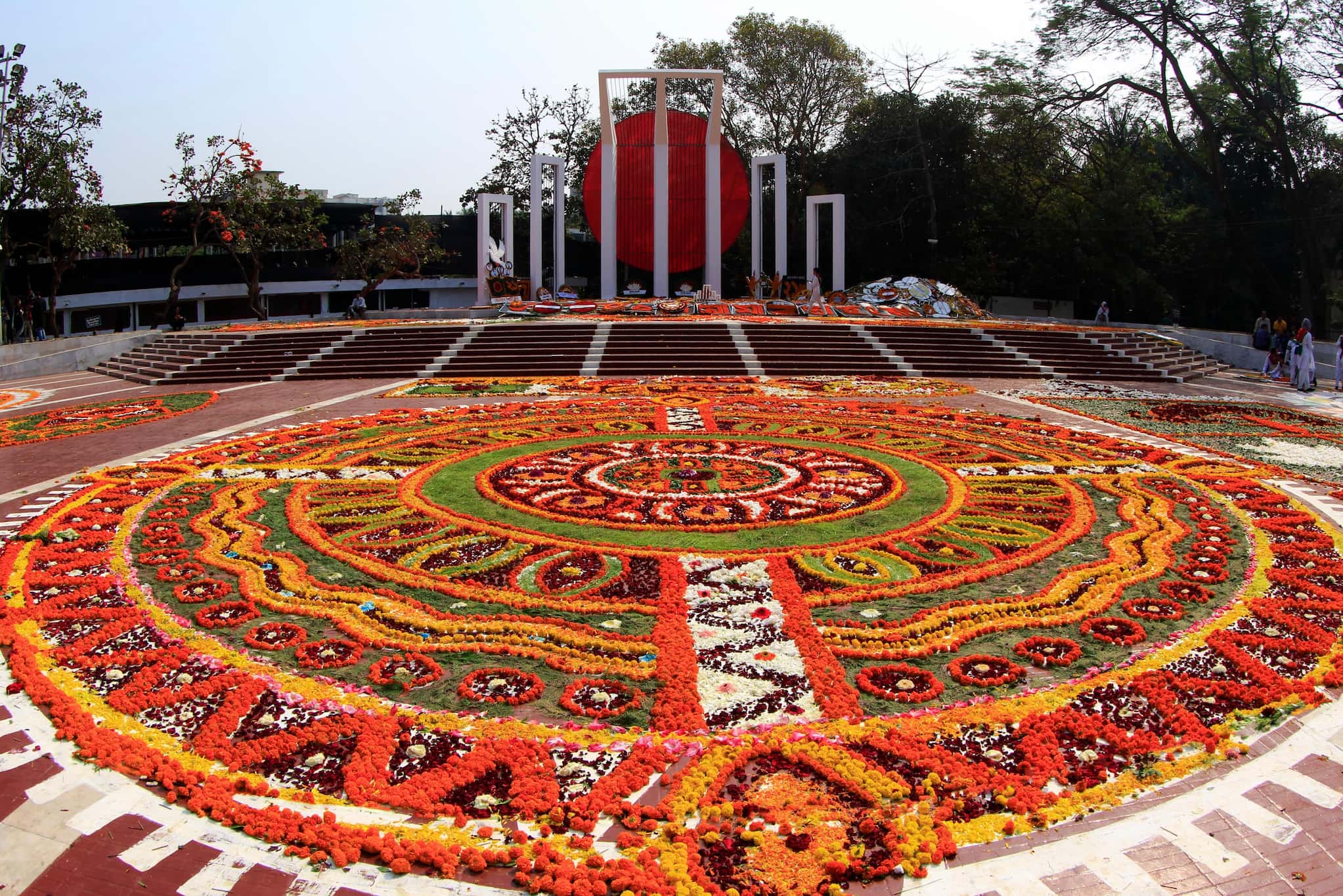 A circular, mosaic-patterned flower bed with reds, yellows, and greens in front of a monument.