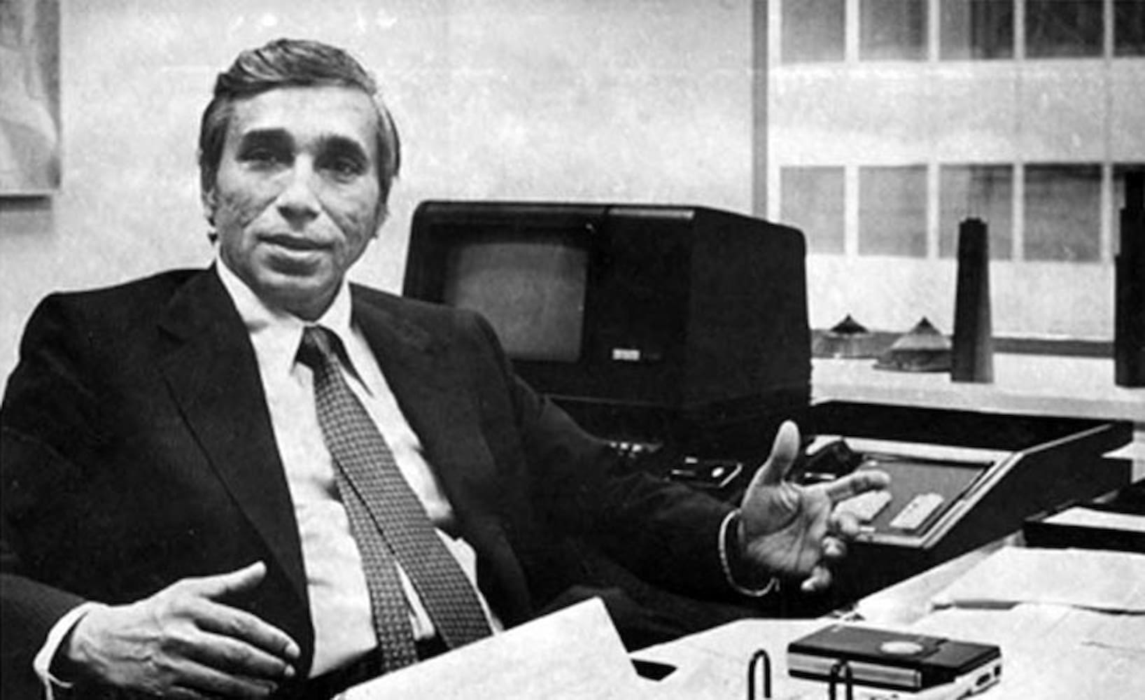 Bangladeshi American Fazlur Khan in a suit and tie sits behind his office desk covered with piles of paper and holds his hands spread apart.