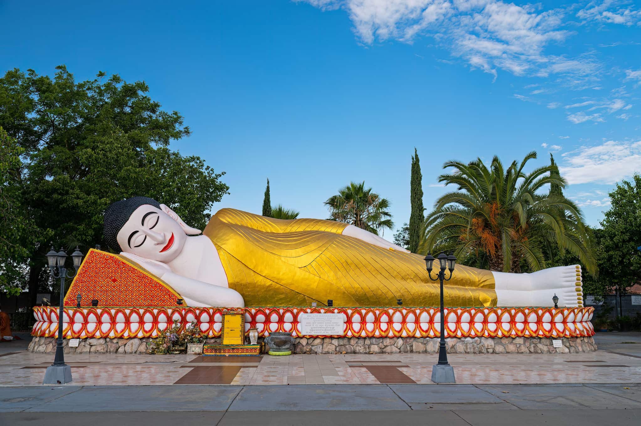 A giant Buddha statue reclines with his head on a pillow and eyes closed. The statue is dressed in a gold robe and rests beneath a cloudy sky.