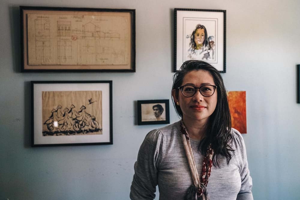 Vietnamese American Kim Luu-Ng in glasses, a colorful scarf, and a long sleeved shirt poses in front of framed images and art honoring her roots.