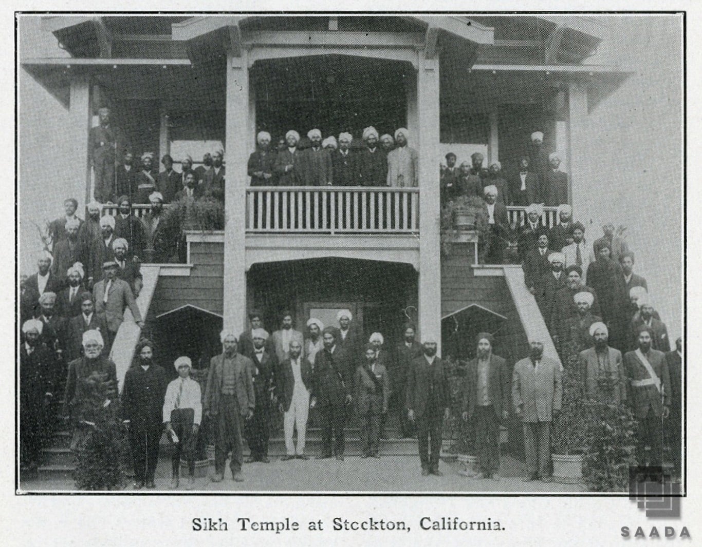 Sikh men wearing turbans, hats, and suits standing along the exterior of the Stockton Gurdwara, a place of Sikh worship and community center.