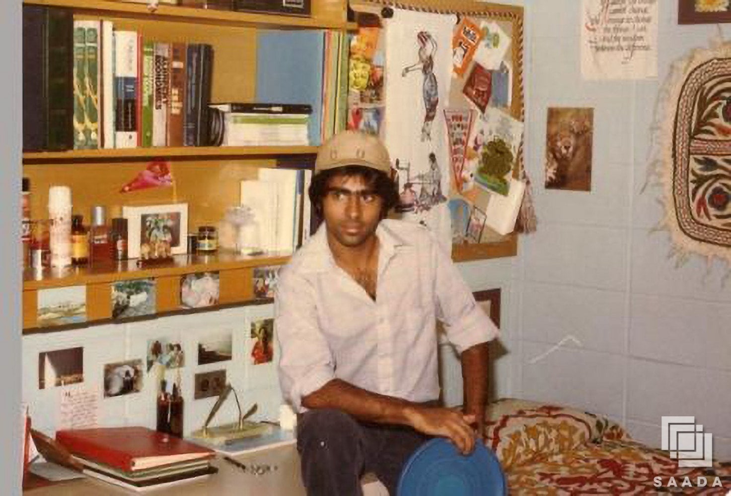 Ali Khataw, wearing a white button down shirt and a tan hat, sits on desk in dorm room. Desk and shelf are stacked with books. Photos cover walls.