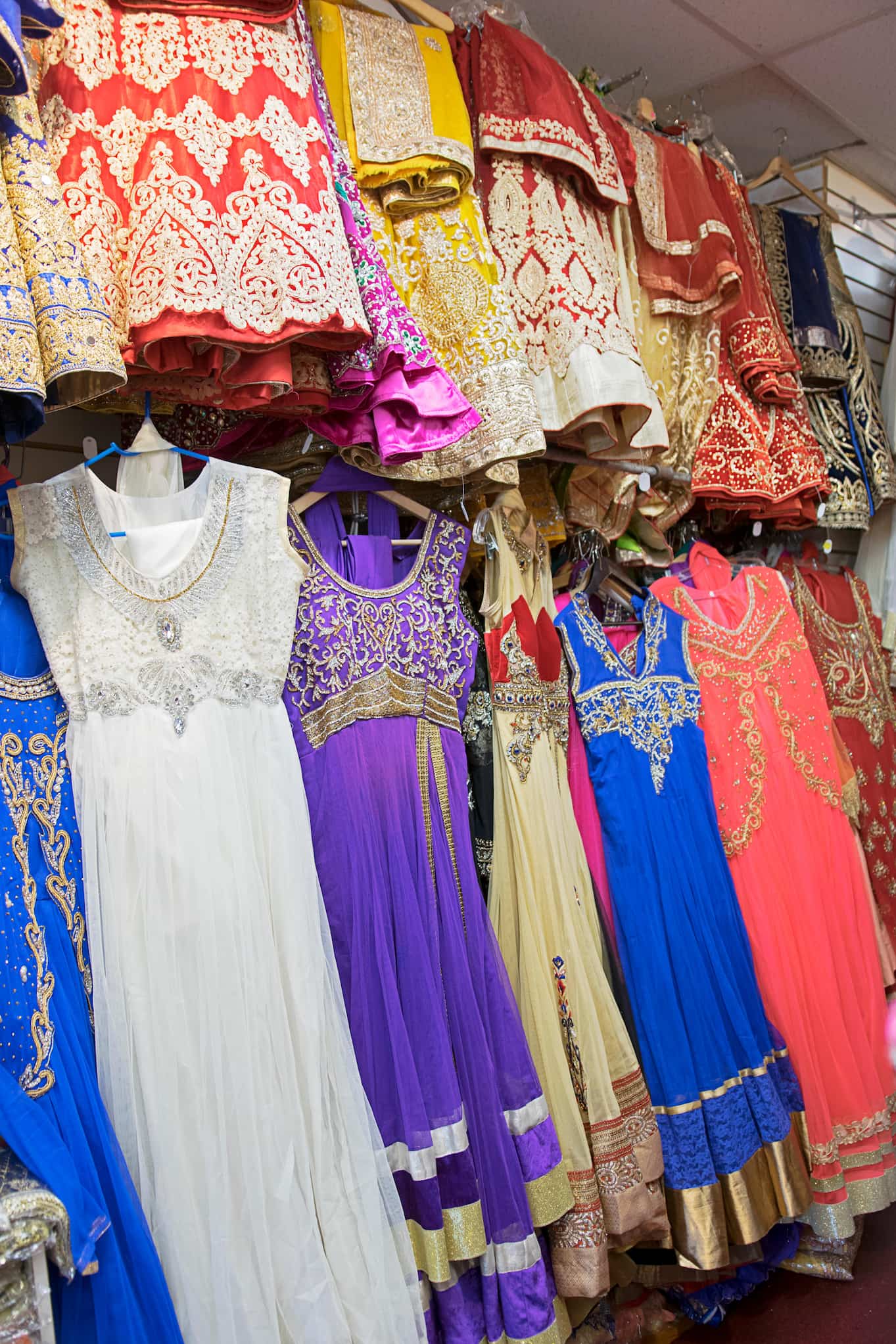 Two rows of colorful shalwar kameez and other garments with intricate embroidered designs hang on display inside a South Asian clothing store.
