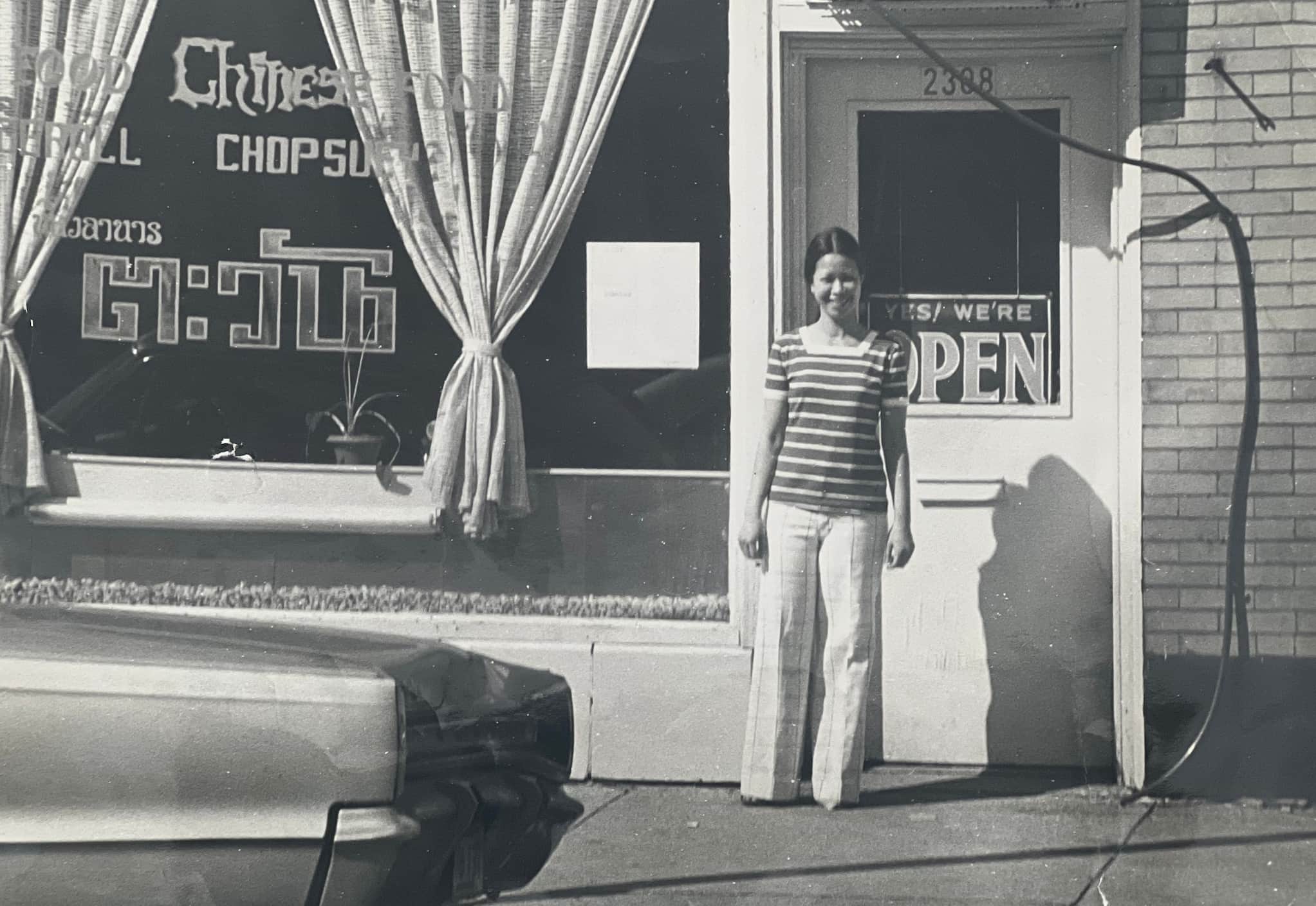 Co-business owner Rushanee Suksod, smiling proudly in a striped shirt, stands by front door of her Thai restaurant Tawan with the open sign displayed.