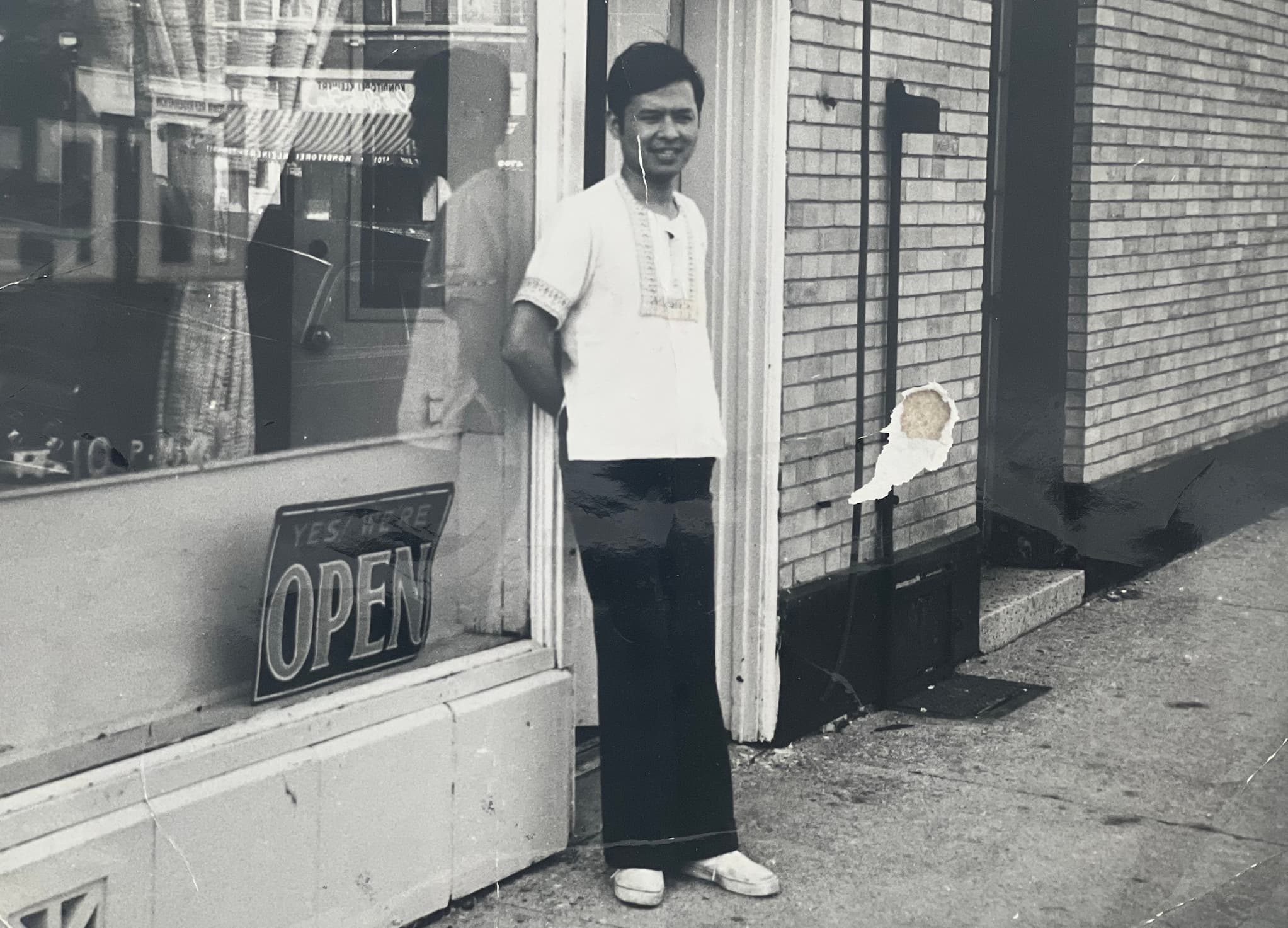 Co-business owner Wanchai Suksod, smiling in black pants and white shirt, leans in doorway of his Thai restaurant Tawan. Open sign shown in window.