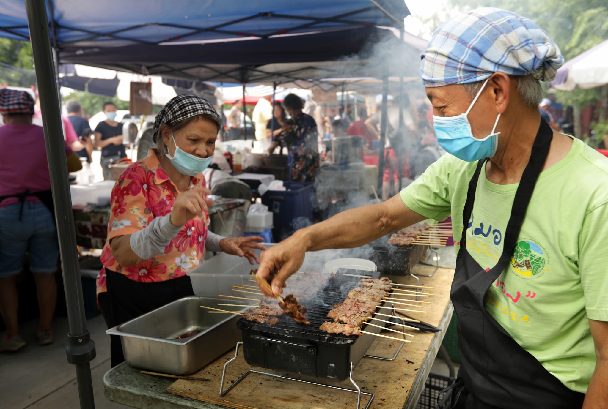 Two food service workers, masked and with hair coverings, prepare skewered meat on grill in outdoor tent. Beside them are more food vendors in tents.