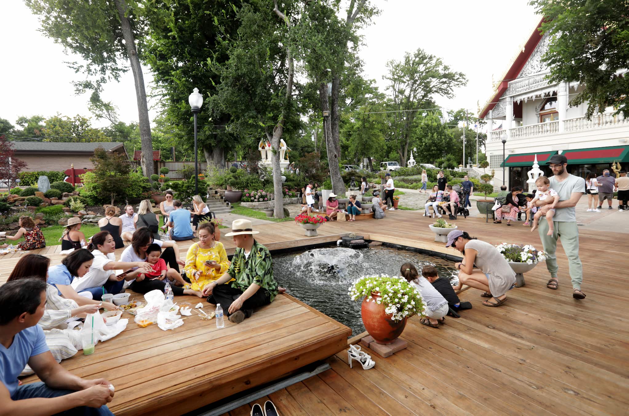 People gather in groups around wooden deck and water feature at outdoor food market. Some sit with food while others walk around landscaped perimeter.