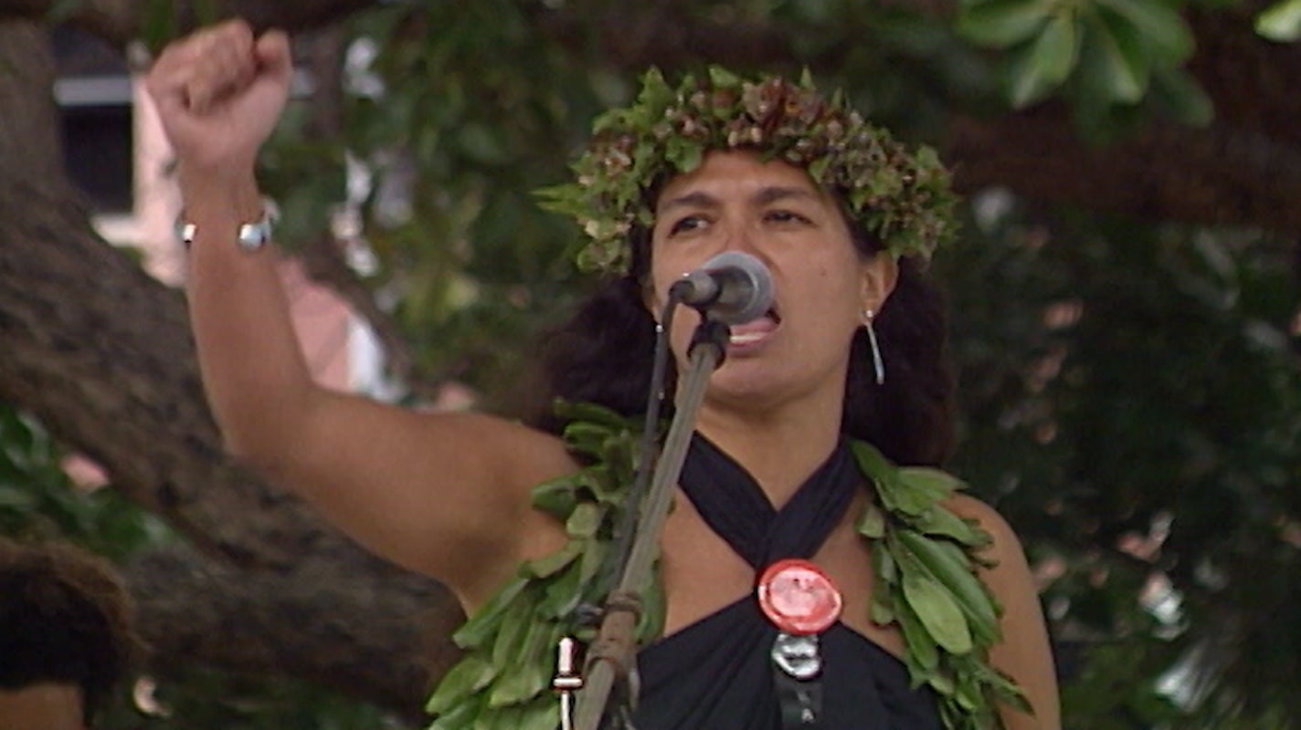 Haunani-Kay Trask, a woman with long, dark hair wearing lei poʻo, speaks passionately into microphone at outdoor event about U.S. imperialism.
