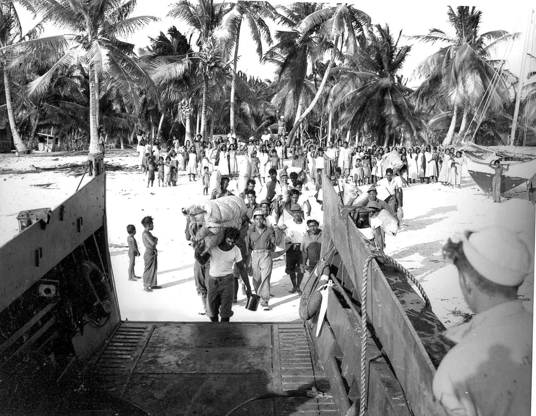 From the point of view of a naval landing craft, a group walks across the beach with their belongings. Behind them, a crowd stands on sand and waves.