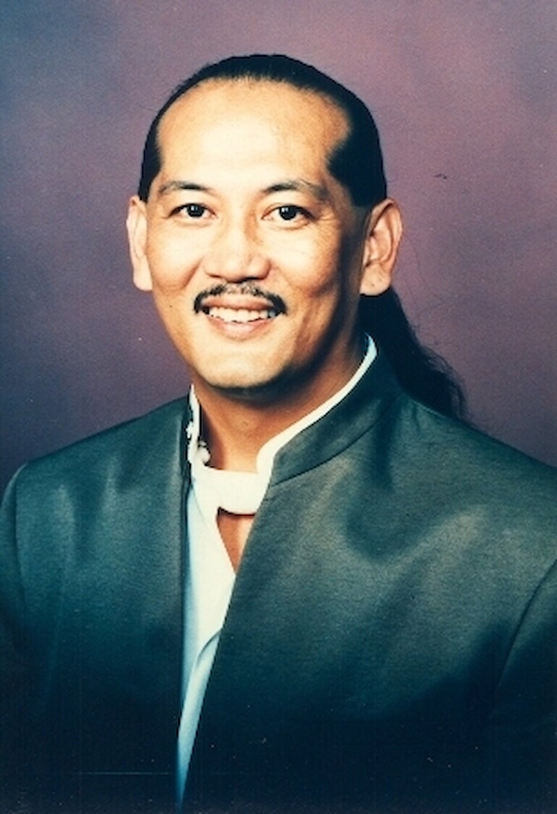 Angel Ånghet LG Santos, a Chamorro rights activist and Guamanian politician with long, black hair, smiles for portrait against purple backdrop.