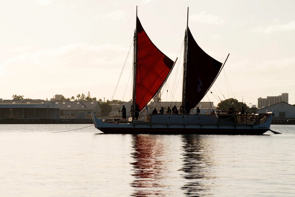Photo of Hōkūleʻa in partial silhouette on the water at Puʻuloa. Its two crab claw sails in crimson red are opened.