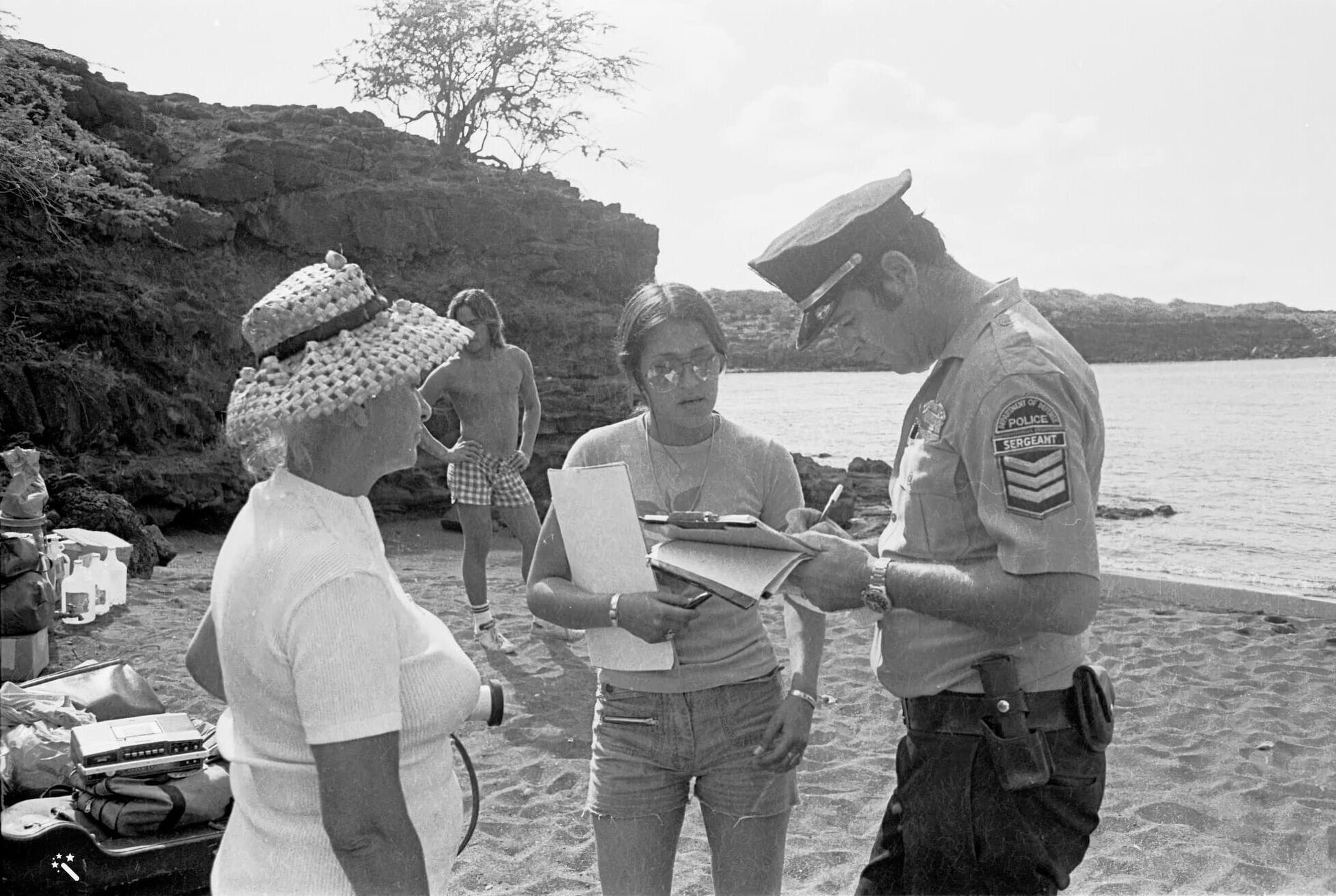 On the shore of Kūheʻeia Bay, a police sergeant writes on a clipboard as two female protestors next to him watch. A shirtless man stands behind them.